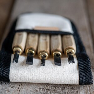 May include: A set of five wood carving chisels with light-coloured wooden handles and brass ferrules, arranged in a white canvas roll-up pouch with black trim. Each tool features a distinct metal blade shape, including V-gouge and flat chisel tips.