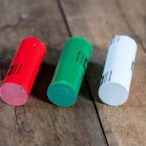 May include: Three cylindrical sticks of wax in red, green, and white. Each stick features a black logo and the text "Made in France" printed on the side. The sticks are arranged in a row, showcasing their solid, smooth, and opaque coloured surfaces.