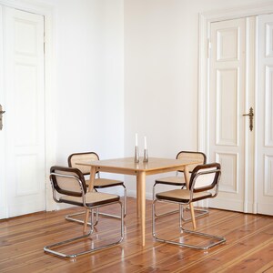 May include: A light wood square dining table with four chairs. The chairs have a woven cane back and seat, with a dark brown frame and chrome legs. Two silver candle holders with white candles are on the table. The room has white doors and wood flooring.