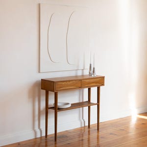Puede incluir: Una consola de madera de estilo moderno de mediados de siglo, con dos cajones y un estante inferior. La mesa tiene patas cónicas y un acabado de madera natural. Dos candelabros plateados y un cuenco blanco están sobre la mesa. Una obra de arte abstracta blanca cuelga de la pared.