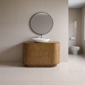 May include: A modern bathroom vanity with a light wood finish, featuring three drawers and a rounded design. It has a white, oval-shaped sink and a circular black-framed mirror. The bathroom also includes a white toilet and light-colored tiled walls and floor.