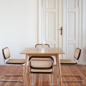 Puede incluir: Una mesa de comedor de madera clara con cuatro sillas. Las sillas tienen asientos y respaldos de mimbre tejido con marcos marrón oscuro y patas cromadas. Dos candelabros plateados están sobre la mesa. La habitación tiene una puerta blanca y suelo de madera.