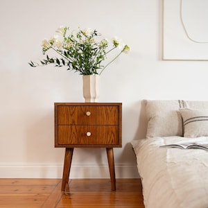 Op de afbeelding: Een houten nachtkastje in mid-century modern stijl met twee lades en messing knoppen. Het nachtkastje bevat een witte vaas met witte bloemen. Het nachtkastje is 61 cm hoog.