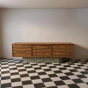 Könnte beinhalten: Ein moderner Holzschrank mit drei Türen, der ein dunkles Holzmaserungsmuster aufweist. Der Schrank steht auf einem zylindrischen Sockel mit einer strukturierten, hellfarbigen Oberfläche. Der Raum hat einen schwarz-weißen Schachbrettboden.