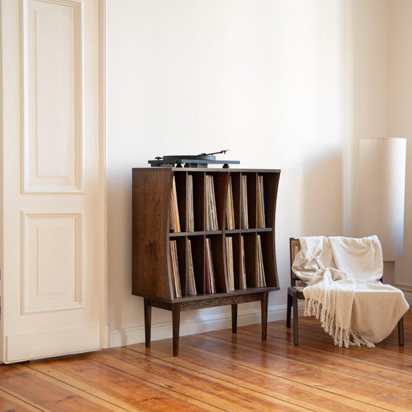 May include: A dark brown wooden record storage cabinet with a turntable on top. The cabinet has slots for vinyl records. A black turntable is on top. A chair with a blanket is next to the cabinet. The cabinet is 100 cm high.
