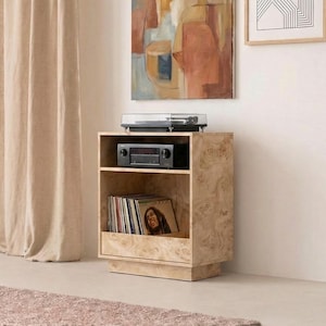 Może przedstawiać: Jasny, drewniany stojak na gramofon z gramofonem, odbiornikiem stereo i płytami winylowymi. Stojak znajduje się w pokoju o neutralnej palecie kolorów, w tym beżowej zasłonie i różowym dywanie. Drewno ma jasne usłojenie.