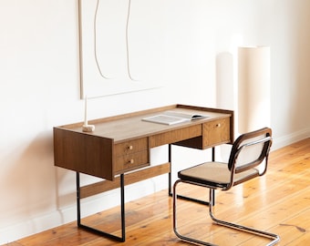 May include: A mid-century modern desk with a light wood finish, two drawers, and black metal legs. A matching chair with a woven seat and chrome frame sits beside the desk. A book lies open on the desk's surface.