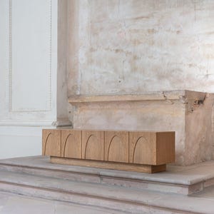 May include: A light brown wooden cabinet with six arched door panels. The cabinet sits on a rectangular base. The background features a textured, off-white wall and a stone ledge.