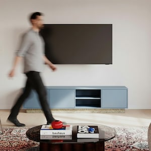 Może przedstawiać: Nowoczesny salon z ciemnym telewizorem na ścianie nad jasnoniebieską szafką. Osoba przechodzi obok okrągłego stolika kawowego z książkami i czerwonym przedmiotem dekoracyjnym. Szafka ma 180 cm szerokości.