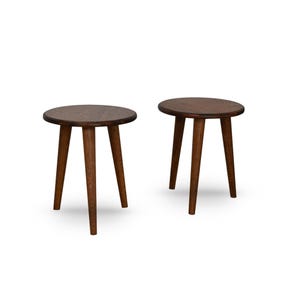 Op de afbeelding: Twee kleine, ronde houten bijzettafels. Elke tafel heeft een donkerbruin tafelblad en drie schuine poten. De tafels zijn gemaakt van donker hout en staan tegen een witte achtergrond.