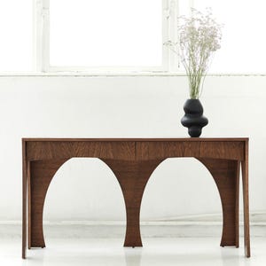 Op de afbeelding: Een donkerhouten console met een uniek gebogen basisontwerp. De tafel heeft een rechthoekig blad en twee lades. Een zwarte, abstracte vaas met gedroogde bloemen staat erop. De tafel staat in een lichte, witte kamer.