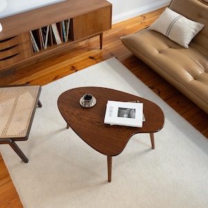 May include: A mid-century modern living room scene featuring a dark brown, organically shaped coffee table with three legs. A small silver cup sits on the table next to a magazine with the word "POINT" visible. A light beige rug and a brown leather sofa are also in the room.