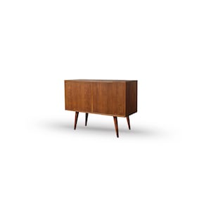 May include: A mid-century modern wooden cabinet with two doors and angled legs. The cabinet is made of a warm-toned wood and has a simple, clean design. The legs are tapered and add to the retro aesthetic.