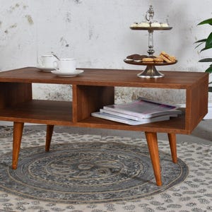 May include: A mid-century modern style coffee table with a warm brown finish. The table has a rectangular top with two open storage compartments and tapered legs. A tiered serving tray with cookies and a plant in a terracotta pot are on the table.