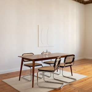 May include: A dining room set featuring a rectangular wooden table with four chairs. The chairs have woven backs and seats, with chrome-finished metal frames. The table is set on a cream-colored rug, with a white wall and artwork in the background.
