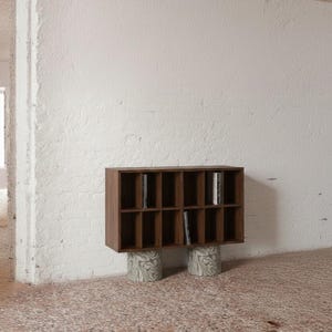 Może przedstawiać: Nowoczesny, brązowy drewniany regał z wieloma przegródkami, spoczywający na dwóch cylindrycznych, wzorzystych podporach. Jednostka jest wypełniona pionowymi przedmiotami, prawdopodobnie książkami lub płytami, na tle białej ściany i nakrapianej podłogi.