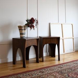 May include: Two dark brown wooden side tables with arched legs. One table holds a decorative vase with a striped pattern and a bouquet of red flowers. The tables are placed on a hardwood floor with a patterned rug.