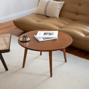 May include: A round, brown wooden coffee table with three angled legs. A small silver cup and saucer sit on the table, along with two books. A tan sofa and a woven chair are in the background.