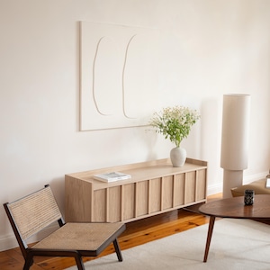 Op de afbeelding: Een moderne woonkamer met een licht houten kast, een stoel met vlechtwerk en een ronde houten salontafel. Een witte vaas bevat bloemen en een hoge, cilindrische lamp staat ernaast. Een wit abstract kunstwerk hangt aan de muur.