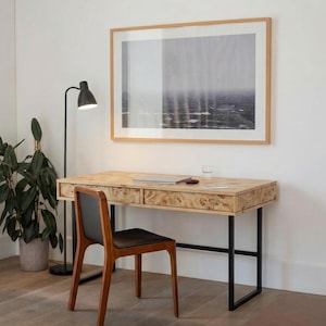Op de afbeelding: Een licht houten bureau met twee lades, een zwart metalen frame en een bijpassende houten stoel met een zwarte zitting. Een zwarte lamp en een plant in een pot staan links. Boven het bureau hangt een ingelijst landschapskunstwerk.