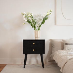 May include: A black nightstand with two drawers and gold knobs. The nightstand has four angled legs. A beige vase with white flowers sits on top. The scene includes a bed with a beige comforter and pillows.