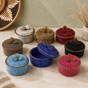 May include: A selection of beaded round boxes in diverse colours, such as blue, red, black, and brown. Each box features a lid with a decorative beaded accent. The boxes are displayed on a neutral surface, with a woven basket and dried plants in the background.