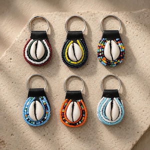 Peut inclure: Six porte-clés composés d'un coquillage cauri blanc central entouré de perles de rocaille colorées sur un support en similicuir noir avec un anneau en métal argenté. Chaque porte-clé présente un motif unique de perles rouges, blanches, vertes, bleues, jaunes et orange.
