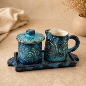 May include: A ceramic set featuring a lidded sugar bowl and a creamer pitcher, both decorated with white hand-painted concentric circles on a mottled blue glaze. The pieces sit on a matching rectangular tray with small side handles.