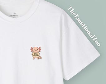 Puede incluir: Camiseta blanca con un pequeño gráfico de ajolote y el texto "Still Evolving". The Emotional Zoo está impreso en la camiseta. El ajolote es de color naranja claro con branquias rosas y una cara sonriente. La camiseta está hecha de un material suave y cómodo.