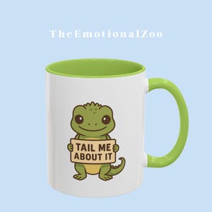 Könnte beinhalten: Eine weiße Keramiktasse mit grünem Rand und Henkel. Die Tasse zeigt eine Comic-Illustration eines grünen Leguan, der ein Schild mit der Aufschrift "Tail Me About It" hält.