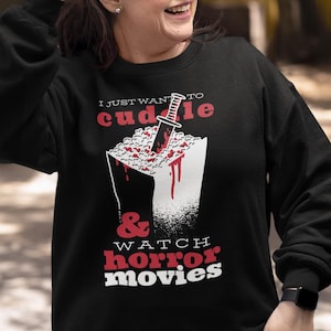 Może przedstawiać: Czarna bluza z napisem "I just want to cuddle & watch horror movies". Grafika przedstawia pudełko popcornu z nożem i kapiącą krwią. Bluza jest noszona z niebieskimi dżinsami.