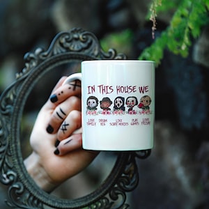 Peut inclure: Mug en céramique blanche avec le texte "IN THIS HOUSE WE" et les phrases "LOVE FAMILY", "DREAM BIG", "LIKE SCARY MOVIES", "PLAY GAMES" et "LOVE FRIDAY". La tasse présente des personnages d'horreur de dessins animés.