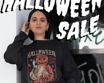 May include: A black long-sleeve sweatshirt featuring a graphic print of a pumpkin-headed figure with skeletal hands wearing a robe. The text on the graphic reads HALLOWEEN IS MY PASSION. The sweatshirt has a crew neckline and long sleeves.