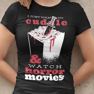 Może przedstawiać: Czarny t-shirt z grafiką przedstawiającą pudełko popcornu z nożem, plamami krwi i napisem "I just want to cuddle & watch horror movies."