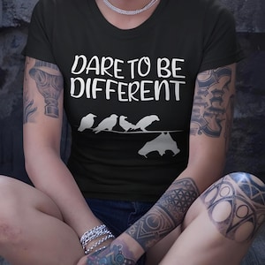 Könnte beinhalten: Schwarzes T-Shirt mit dem weißen Text "DARE TO BE DIFFERENT", zusammen mit einer Grafik von Vögeln auf einem Draht und einer Fledermaus. Das T-Shirt hat einen Rundhalsausschnitt.