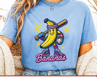 May include: Pale blue t-shirt with a cartoon banana character holding a baseball bat and ball. The banana is yellow with a smiling face, wearing a headband, and athletic shoes. The word "Bananas" is written in pink script below the character.