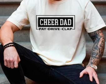 May include: Cream-colored t-shirt with the text "CHEER DAD" in a black rectangular box. Below the text are the words "PAY • DRIVE • CLAP" in a smaller font. The shirt is made of a soft material.