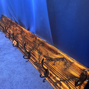 Puede incluir: Perchero de madera con diseño de madera quemada y ganchos de metal negro. El perchero es de madera marrón claro con detalles en marrón oscuro y negro. Los ganchos están espaciados uniformemente.