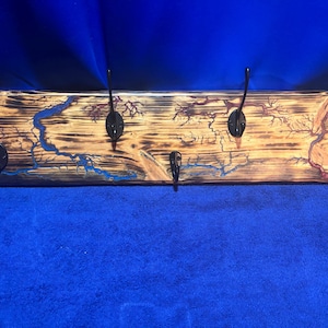Op de afbeelding: Een houten kapstok met vijf zwarte metalen haken. Het hout heeft een verbrande afwerking met blauwe en rode accenten, die lijken op een kaart of bliksem. De kapstok is rechthoekig.