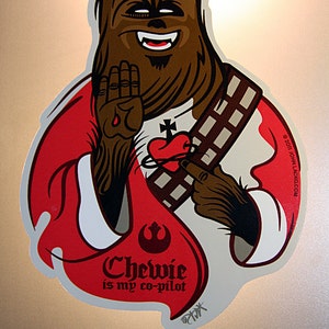 May include: A red and white sticker featuring a cartoon illustration of Chewbacca from Star Wars. Chewbacca is wearing a red robe and a white shirt with a heart and cross on it. The text "Chewie is my co-pilot" is printed on the sticker.