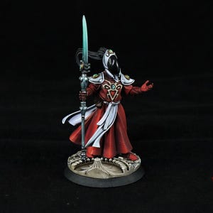 Könnte beinhalten: Eine bemalte Miniaturfigur eines Kriegers in rotem Gewand mit weißen Akzenten, schwarzem Helm und silberner Schulterpanzerung. Die Figur hält einen hohen, schlanken Speer mit leuchtend türkisfarbener Klinge und steht auf einem runden Steinsockel.