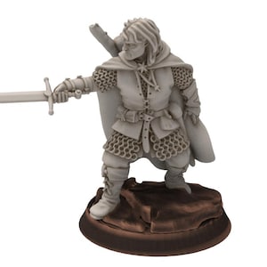 Puede incluir: Una figura en miniatura de un guerrero que lleva una armadura de cota de malla y una capa con capucha, sosteniendo una espada y de pie sobre una base rocosa.