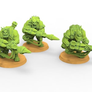Peut inclure: Trois figurines miniatures vertes représentant des créatures ressemblant à des Orcs, chacune armée d'une grande arme futuriste. Elles sont en position accroupie sur de petites bases circulaires de couleur beige. Les figurines ont des armures et des traits faciaux détaillés.