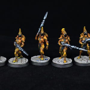 Könnte beinhalten: Fünf Miniatur-Kriegerfiguren in gelber Rüstung mit schwarzen Helmen, die jeweils eine lange, metallisch blaue Waffe halten. Sie stehen auf runden Sockeln mit steinartiger Textur. Ein rotes Logo in der Ecke zeigt CREED'S PAINTING.