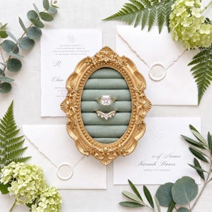 May include: An oval gold ornate frame containing a sage green velvet ring insert with horizontal slots. Two rings are displayed: one with a large square-cut stone and one with a V-shaped band featuring small stones, surrounded by wedding stationery.
