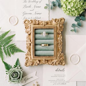 May include: A rectangular gold ornate frame containing a sage green velvet ring holder with five slots. Two gold rings are placed in the slots, one with a marquise-cut stone. The frame is surrounded by wedding stationery, white envelopes with wax seals, and green floral accents.