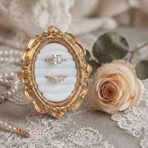 May include: An oval ring holder featuring an ornate gold-toned frame with intricate floral carvings. The center is filled with white velvet padding containing horizontal slots that hold two rings: one solitaire ring and one V-shaped band.