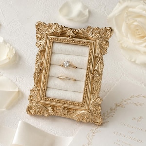 Może przedstawiać: Złoty, prostokątny stojak na pierścionki z ozdobną, rzeźbioną ramą i białym aksamitnym wnętrzem z pięcioma poziomymi szczelinami. W szczelinach umieszczono dwa pierścionki: jeden z pojedynczym okrągłym kamieniem i jeden z małymi kamieniami.