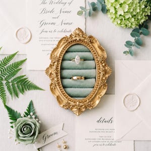 May include: An oval gold-toned ornate frame containing a sage green velvet ring holder with two rings, one marquise-cut diamond ring and one plain gold band, displayed alongside wedding stationery, a green rose boutonniere, and greenery.