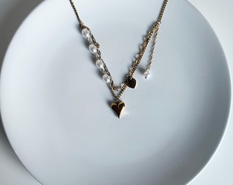May include: Gold-toned necklace with a heart pendant and pearl accents. The necklace features multiple strands, with a heart-shaped charm at the center and small pearls along one strand. A delicate piece of jewelry.